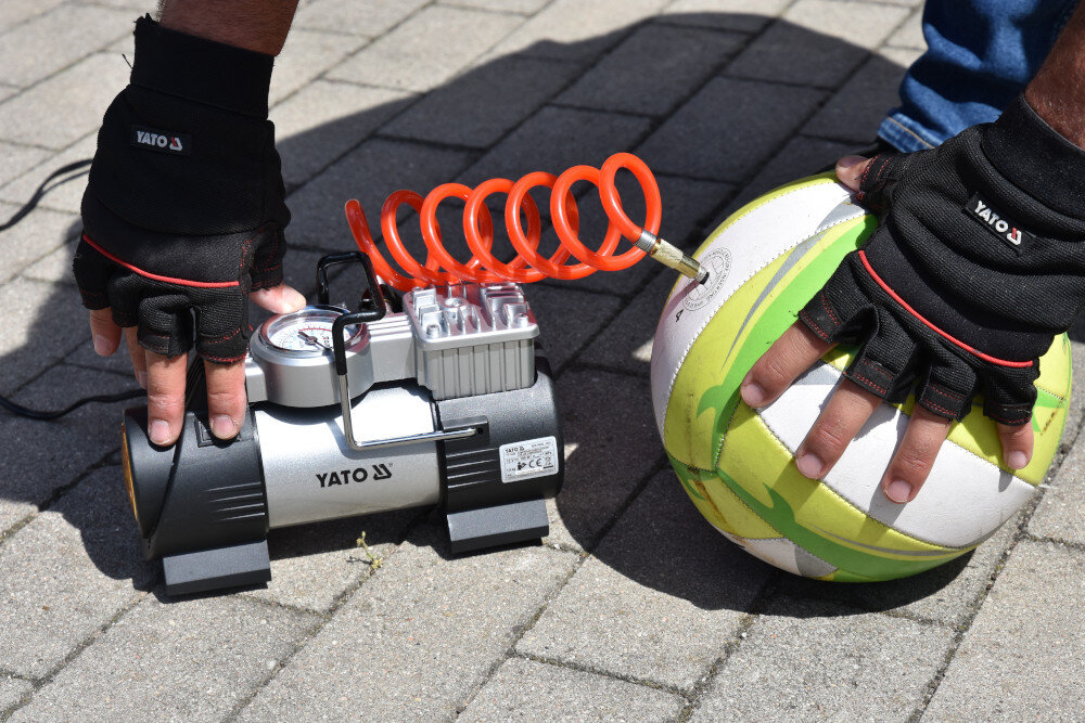 Na zdjęciu widać kompresor samochodowy z logo YATO, podłączony do piłki za pomocą pomarańczowego węża. Osoba w rękawiczkach trzyma piłkę i obsługuje kompresor na chodniku z kostki brukowej.