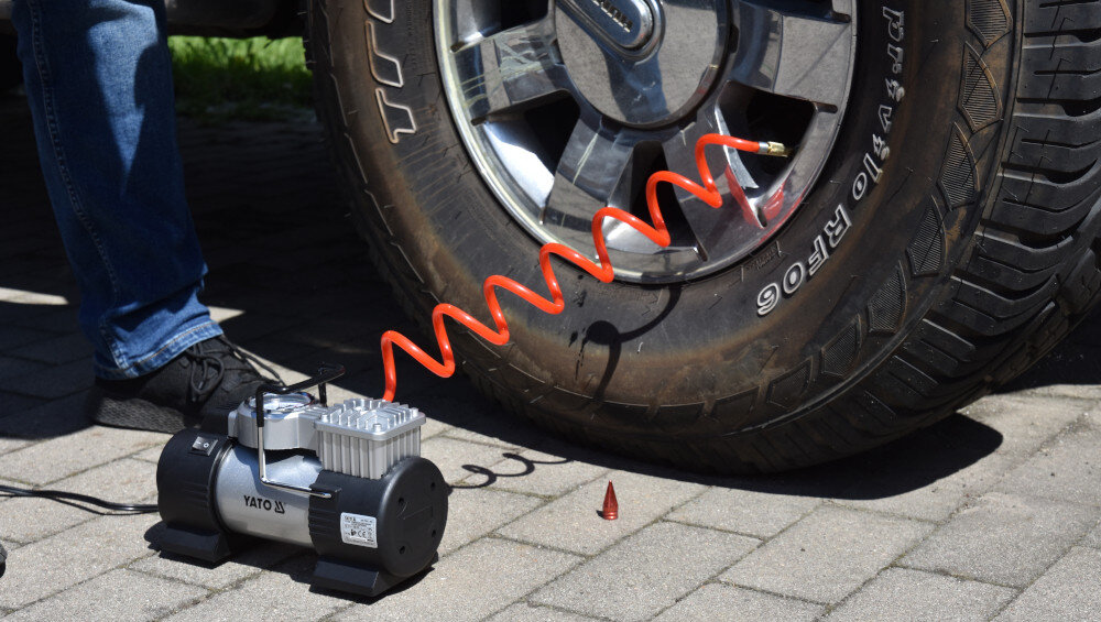 Na obrazie widoczny jest kompresor samochodowy z napisem \'YATO\' podłączony do opony samochodowej za pomocą spiralnego, pomarańczowego przewodu. Obok kompresora leży mała, czerwona nasadka.
