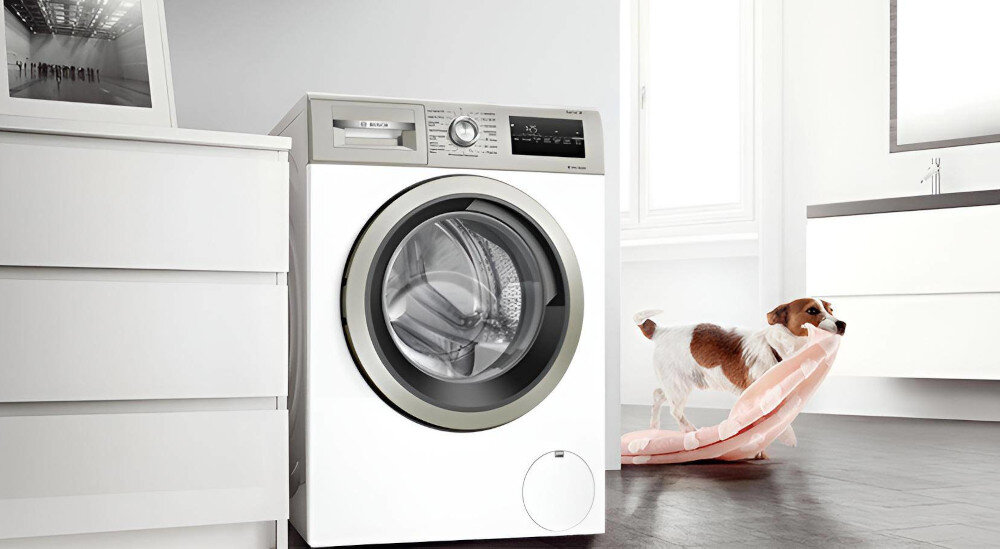 Pralka o białej obudowie z widocznym panelem sterowania i okrągłym bębnem. W tle jasne wnętrze z psem trzymającym różowy koc.