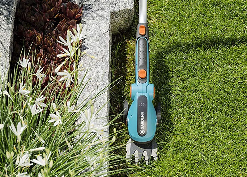 Na obrazie widoczne są nożyce do trawy z logo \'GARDENA\', umieszczone na trawniku obok kamiennej krawędzi i białych kwiatów. Nożyce mają turkusowy kolor z pomarańczowymi elementami.