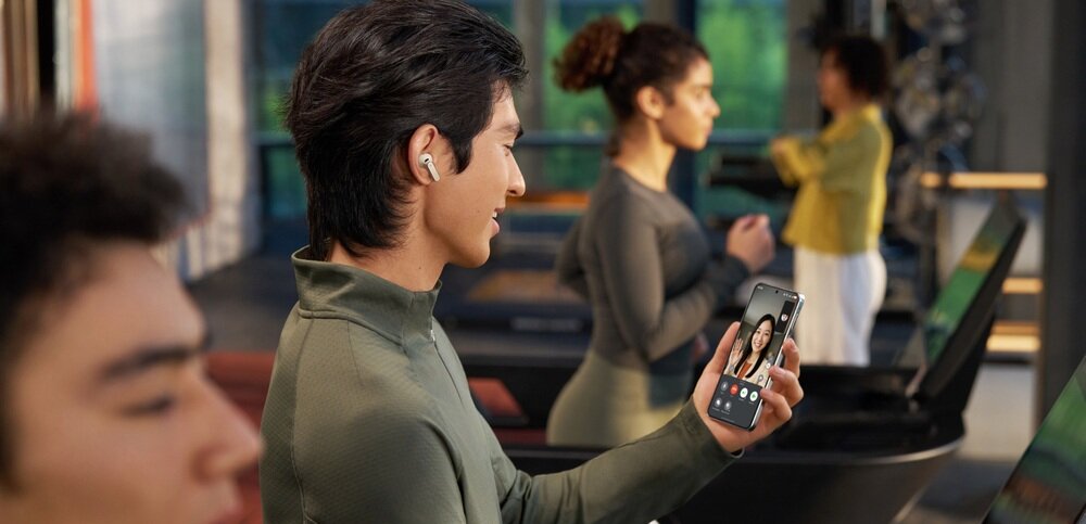 Osoba trzymająca telefon z widocznym ekranem, na którym odbywa się wideorozmowa. W uchu ma słuchawkę bezprzewodową, a w tle widać inne osoby na bieżniach.