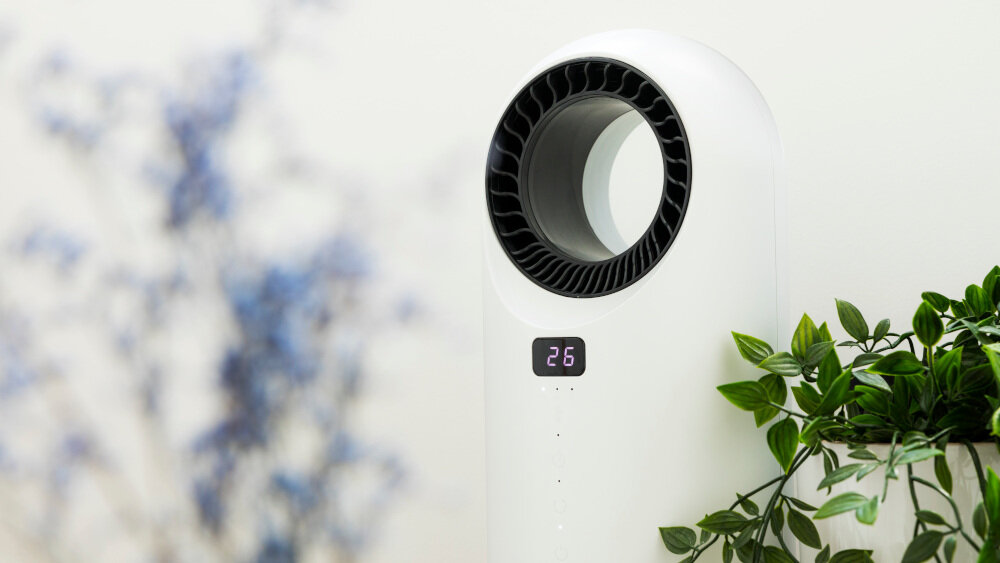 Biały termowentylator bezłopatkowy z wyświetlaczem pokazującym liczbę 26, otoczony roślinami. W tle widoczne są rozmyte niebieskie i zielone elementy dekoracyjne.