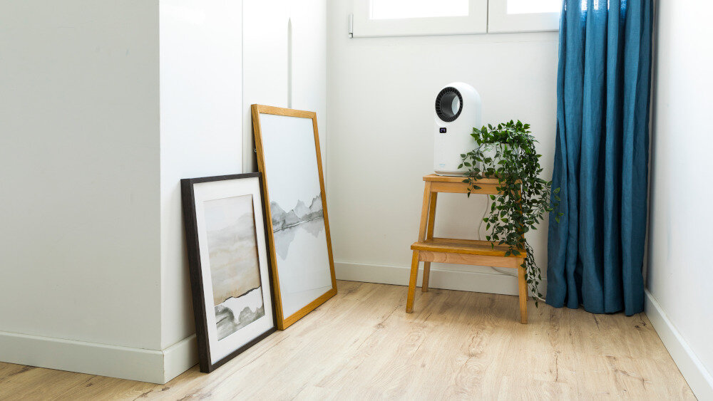 W jasnym pomieszczeniu na drewnianym stołku stoi biały termowentylator o nowoczesnym designie, obok niego znajduje się roślina doniczkowa. Na podłodze oparto o ścianę dwa obrazy w ramach, a w tle widać niebieską zasłonę i okno.