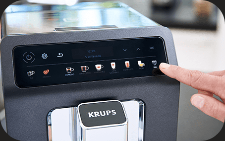 Ekspres do kawy z panelem dotykowym, na którym widoczne są ikony różnych rodzajów kawy oraz logo KRUPS. Palec wskazuje na jedną z ikon na panelu.