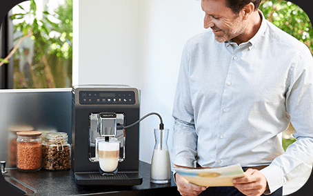 Na blacie kuchennym stoi ekspres do kawy z filiżanką pod dyszą, obok znajduje się karafka z mlekiem. W tle widać osobę trzymającą kartkę papieru.