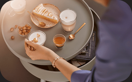 Na okrągłym stoliku znajdują się filiżanki z kawą, talerz z ciastem i orzechy. Obok leży książka i złota łyżeczka.