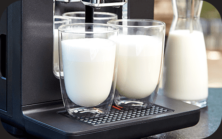 Na obrazie widoczny jest ekspres do kawy z dwoma szklankami wypełnionymi mlekiem. Obok znajduje się karafka z mlekiem.