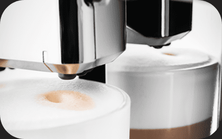 Zbliżenie na dwa szklane kubki z mleczną pianką, pod którymi znajduje się dysza ekspresu do kawy. Kubki są częściowo wypełnione kawą z widoczną warstwą mleka.