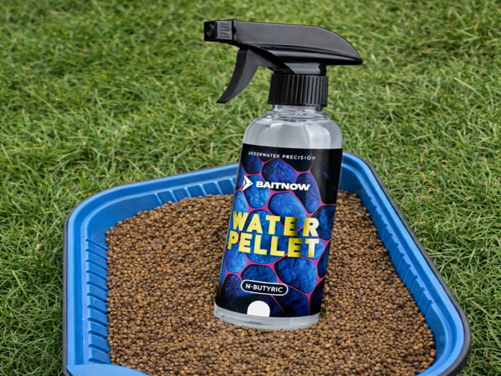 Butelka z rozpylaczem o nazwie \'BAITNOW WATER PELLET N-BUTYRIC\' stoi na niebieskiej tacy wypełnionej granulatem, na tle trawy. Na etykiecie widnieje napis \'UNDERWATER PRECISION\'.