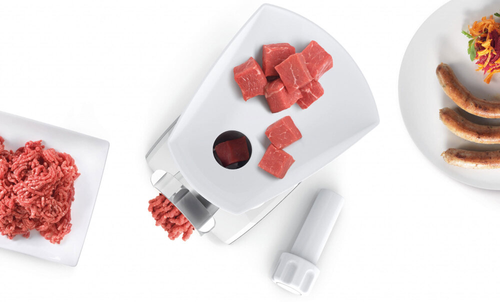 Na obrazie widoczna jest maszynka do mięsa z kawałkami surowego mięsa na tacy oraz zmielonym mięsem na talerzu. Obok znajduje się talerz z kiełbasami i surówką.