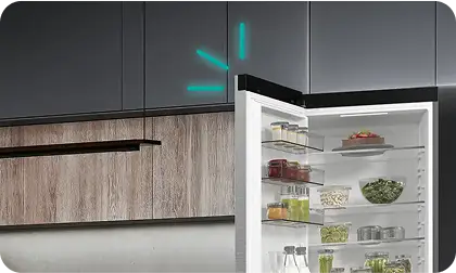 Otwarta lodówka z widocznymi półkami wypełnionymi różnymi produktami spożywczymi, takimi jak warzywa i słoiki. W tle widoczne są szafki kuchenne w ciemnym kolorze.