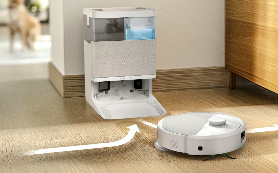 Na podłodze znajduje się biały robot sprzątający obok stacji dokującej z otwartą klapą. W tle widoczny jest rozmyty obraz psa.