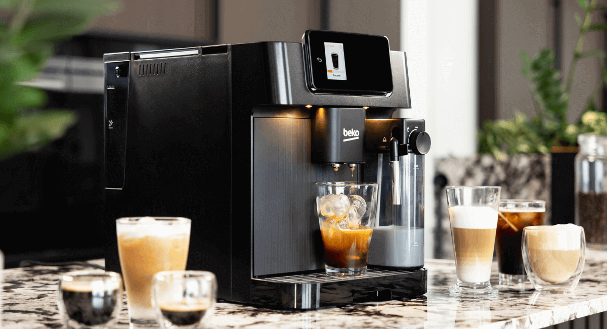 Na marmurowym blacie stoi czarny ekspres do kawy z logo \'beko\', obok którego znajdują się różne szklanki z kawą. W tle widoczne są rośliny i rozmyte elementy wnętrza.