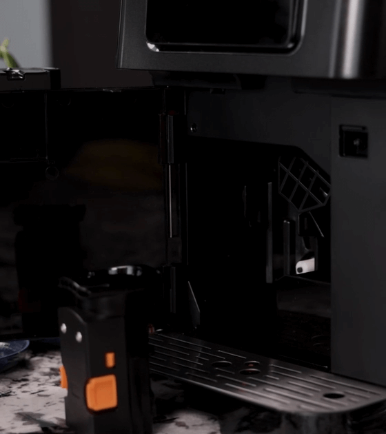 Otwarty ekspres do kawy z widocznym wnętrzem i wyjmowanym pojemnikiem. Na blacie znajduje się tacka ociekowa z otworami.