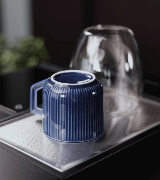 Na metalowej tacy stoją do góry dnem niebieski kubek z uchwytem i przezroczysta szklanka. W tle widoczne są rozmyte zielone liście rośliny.