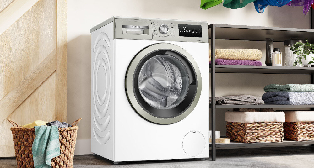 Pralka o białej obudowie z widocznym panelem sterowania i okrągłym bębnem. Obok znajduje się półka z ręcznikami i kosz z praniem.