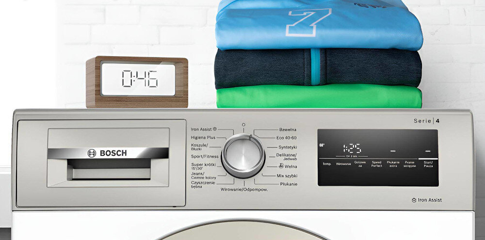 Pralka z panelem sterowania, na którym widoczne są różne programy prania oraz wyświetlacz pokazujący czas 1:25. Na pralce leży stos złożonych ubrań i zegar elektroniczny pokazujący 0:46.