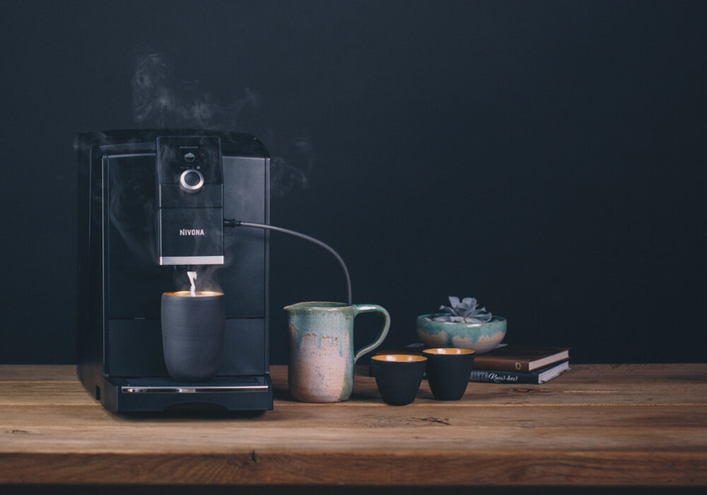 Ekspres do kawy z napisem NIVONA na drewnianym blacie. Obok znajdują się filiżanki, dzbanek i książki.