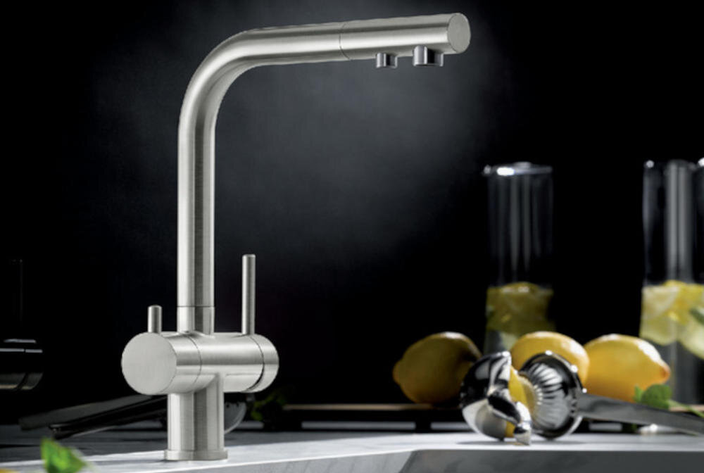 Na obrazie widoczna jest stalowa bateria kuchenna o nowoczesnym designie, zamontowana na blacie. W tle znajdują się cytryny i metalowy wyciskacz do cytrusów.