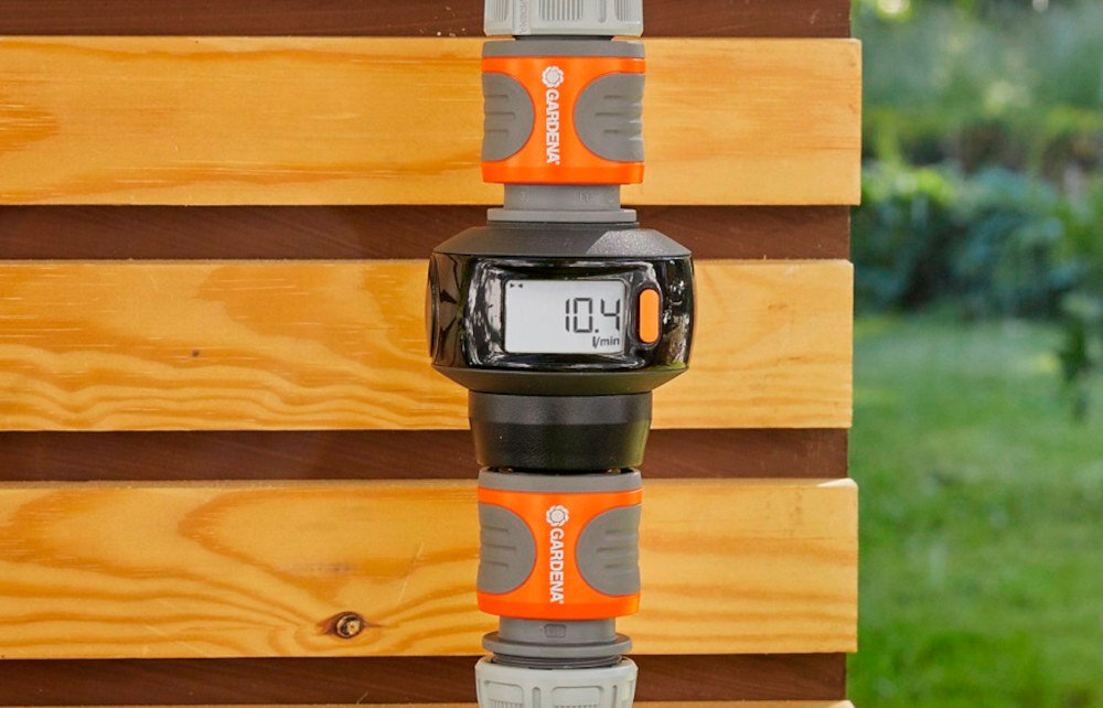 Na obrazie widoczny jest element systemu nawadniania z wyświetlaczem pokazującym \'10.4 l/min\'. Elementy są w kolorze pomarańczowym i szarym z logo \'GARDENA\'.