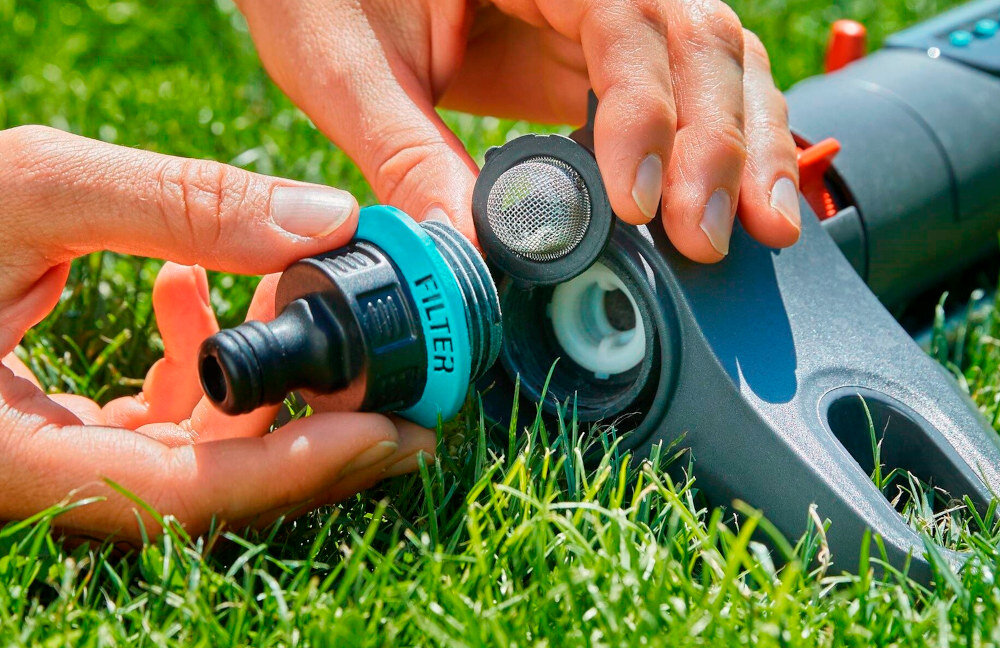 Osoba trzyma element z napisem \'FILTER\' i montuje go do zraszacza ogrodowego na trawie. W tle widoczna jest część zraszacza z otworem i siatką filtrującą.