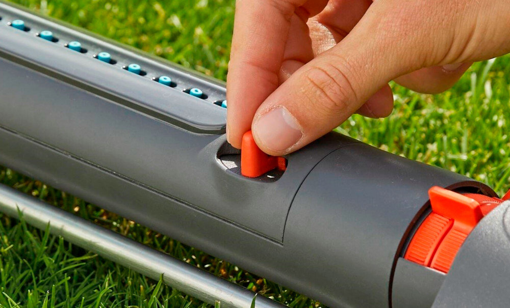Osoba reguluje pomarańczowy element na szarym zraszaczu ogrodowym umieszczonym na trawie. Zraszacz posiada rząd niebieskich dysz na górnej powierzchni.
