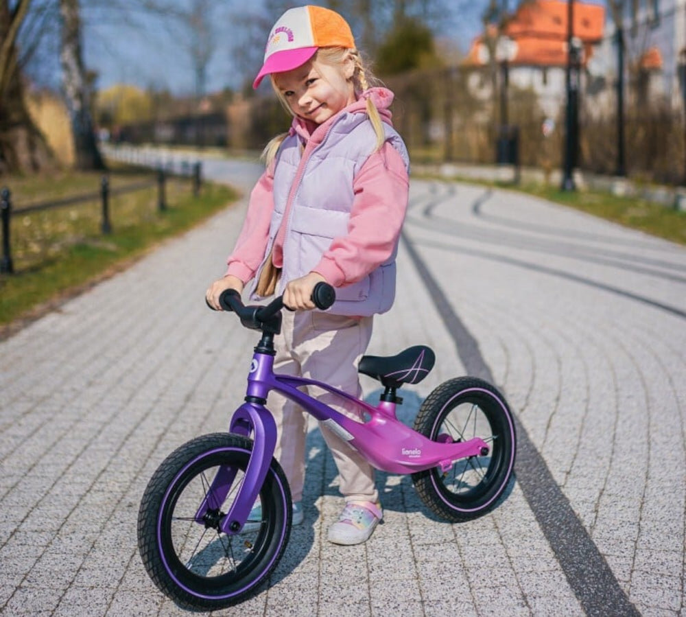 Rowerek biegowy LIONELO Bart Air Różowo-fioletowy park drzewa droga dziecko stoi obok rowerka trzymając go za kierownicę radość rozwój samodzielne przemieszczanie się pierwszy pojazd