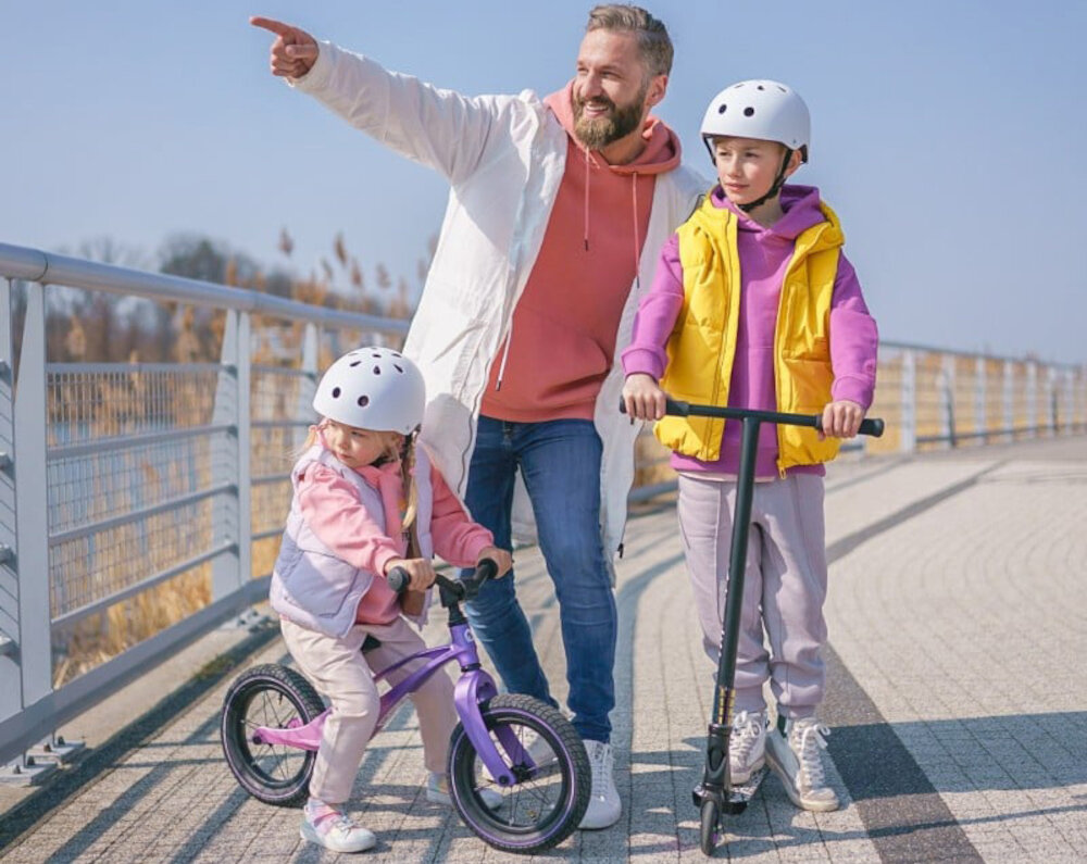 Rowerek biegowy LIONELO Bart Air Różowo-fioletowy jezioro pomost na pomoście dziecko na roweku obok dziecko na hulajnodze obok dorosły mężczyzna certyfikat TÜV spełniania europejskie normy bezpieczeństwa materiały i konstrukcja zostały przetestowane pod kątem wytrzymałości i funkcjonalności odpowiedni do użytku przez dzieci wspiera rozwój psychoruchowy