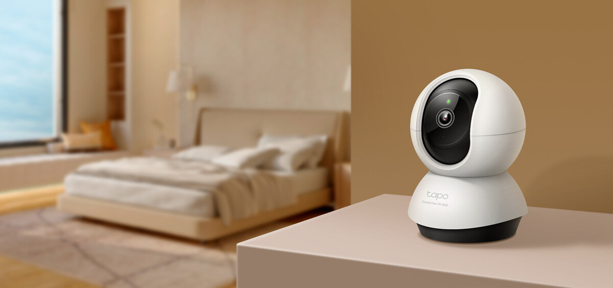 Na pierwszym planie znajduje się biała, obrotowa kamera z logo \'tapo\' na podstawie. W tle widoczna jest sypialnia z łóżkiem i lampką nocną.