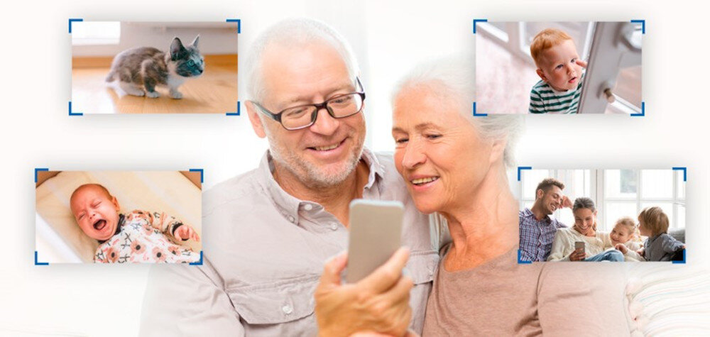 Starsza para ogląda coś na smartfonie, otoczona zdjęciami dzieci i kota w niebieskich ramkach. W tle widać rodzinę siedzącą na kanapie, również patrzącą na urządzenie.