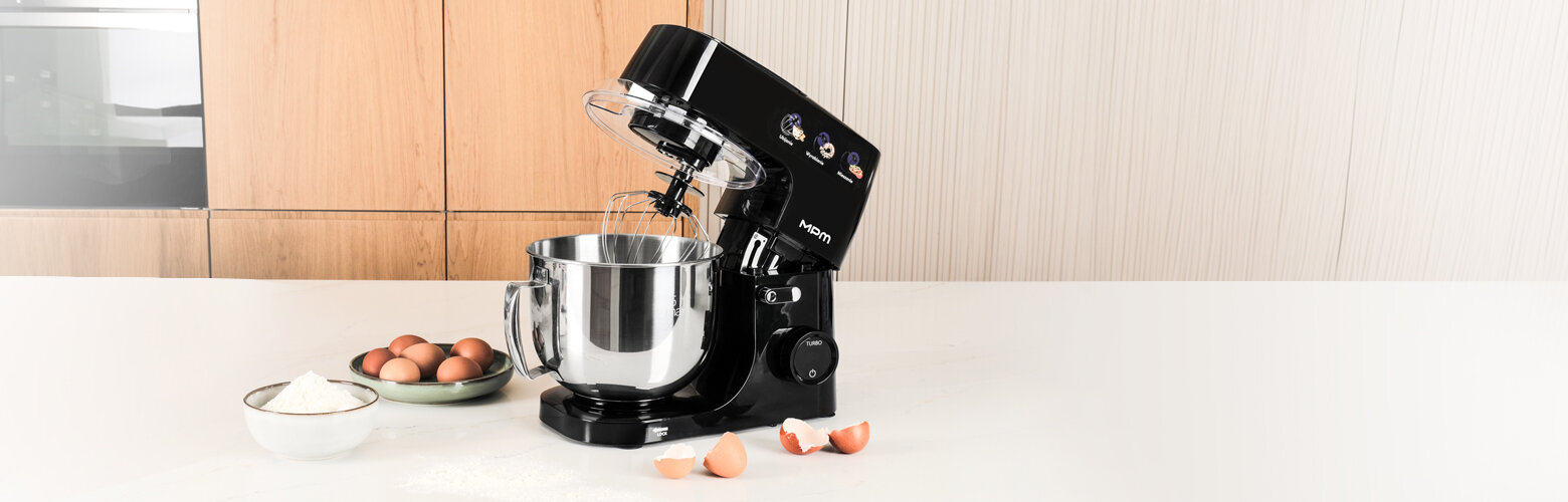 Na blacie kuchennym stoi czarny robot kuchenny z metalową misą. Obok znajdują się jajka i miska z mąką.