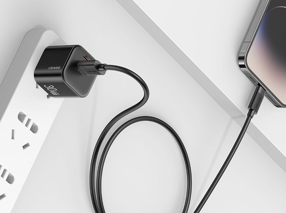Czarny kabel USB-A podłączony do adaptera z logo USAMS, wpięty do listwy zasilającej. Drugi koniec kabla jest podłączony do smartfona.