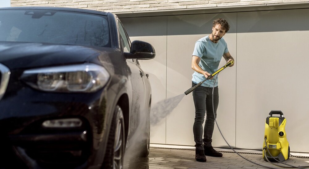 Osoba używa żółtej myjki ciśnieniowej do mycia czarnego samochodu. Myjka stoi na podłożu obok osoby.