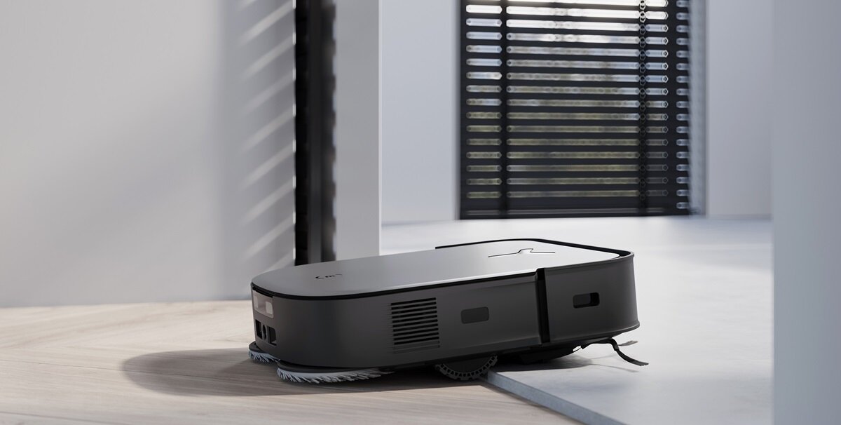 Czarny robot sprzątający porusza się po jasnej podłodze w nowoczesnym wnętrzu. W tle widoczne są pionowe żaluzje na oknie.