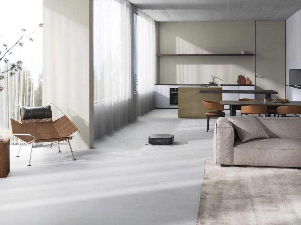 W nowoczesnym salonie z dużymi oknami i minimalistycznym wystrojem znajduje się czarny robot sprzątający na podłodze. W tle widoczna jest kuchnia z wyspą i stołem z krzesłami.