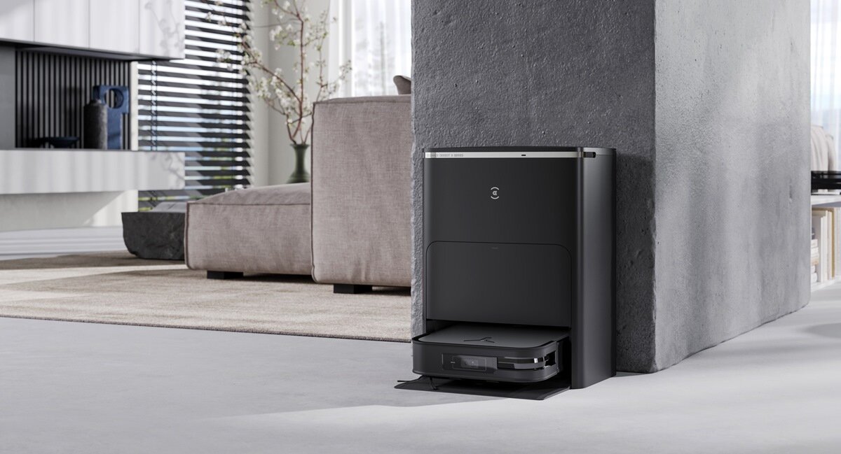 Czarny robot sprzątający znajduje się w stacji dokującej przy ścianie w nowoczesnym wnętrzu. W tle widoczna jest kanapa i okno z żaluzjami.