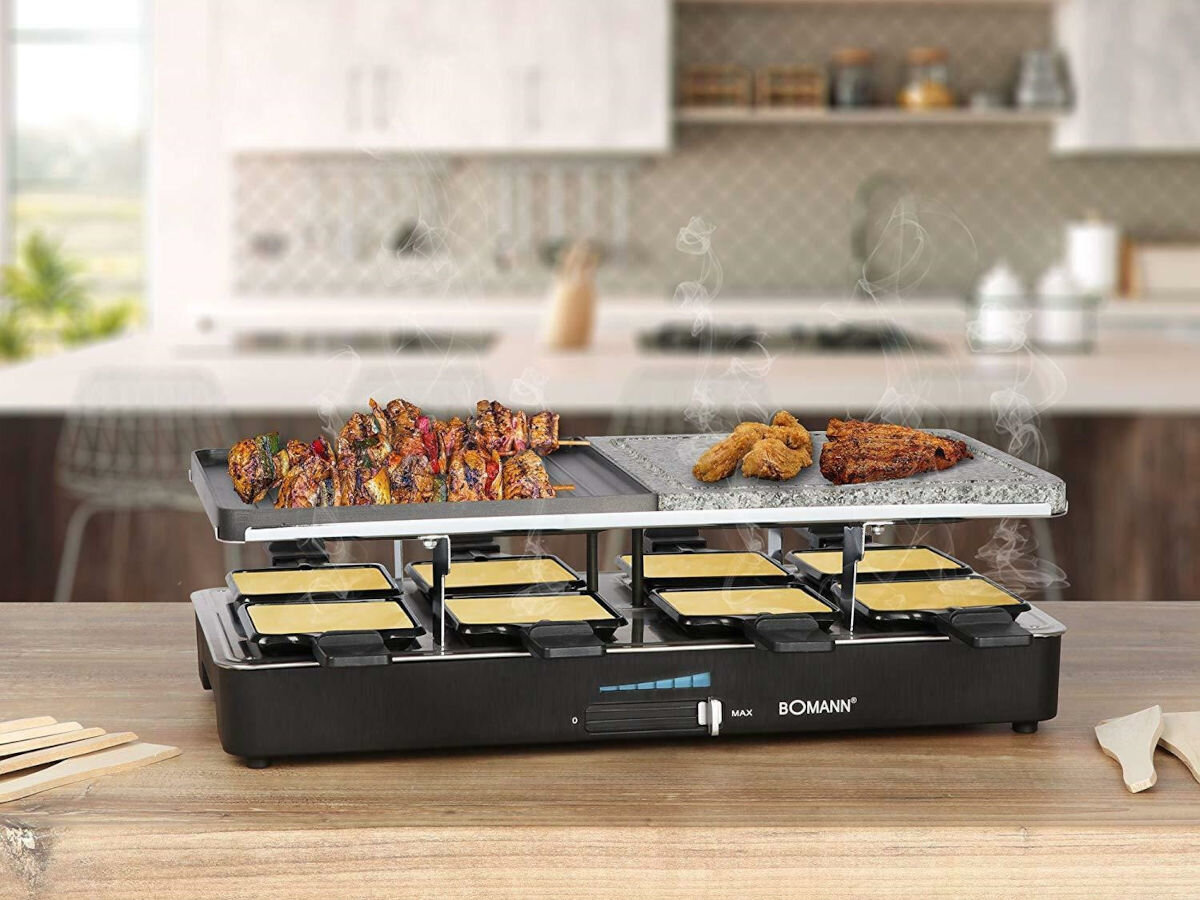 Grill elektryczny z logo \'BOMANN\' na blacie kuchennym, z potrawami na górnej płycie i małymi patelniami poniżej. W tle widoczna jest nowoczesna kuchnia z wyspą i sprzętem kuchennym.