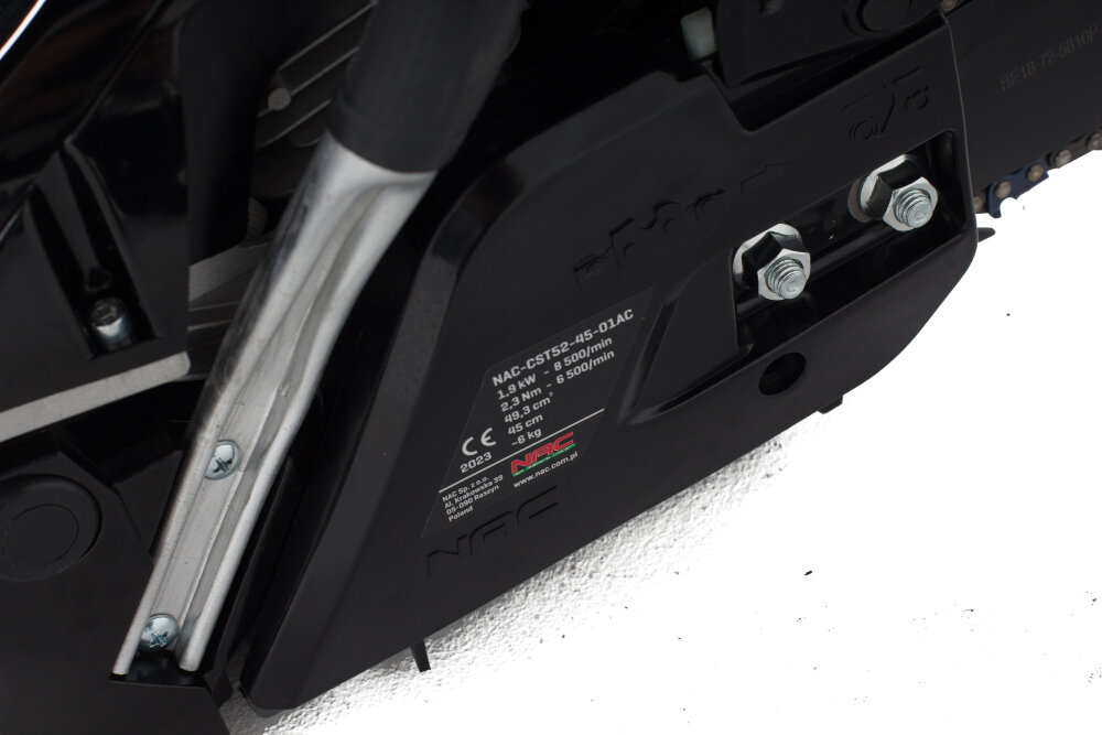 Zbliżenie na część piły spalinowej z widoczną tabliczką znamionową. Na tabliczce widnieje tekst: \'NAC-CST52-45-01AC, 2.3 kW, 3.1 KM, 8500/min, 52 cm3, 45 cm, 0.325\'.