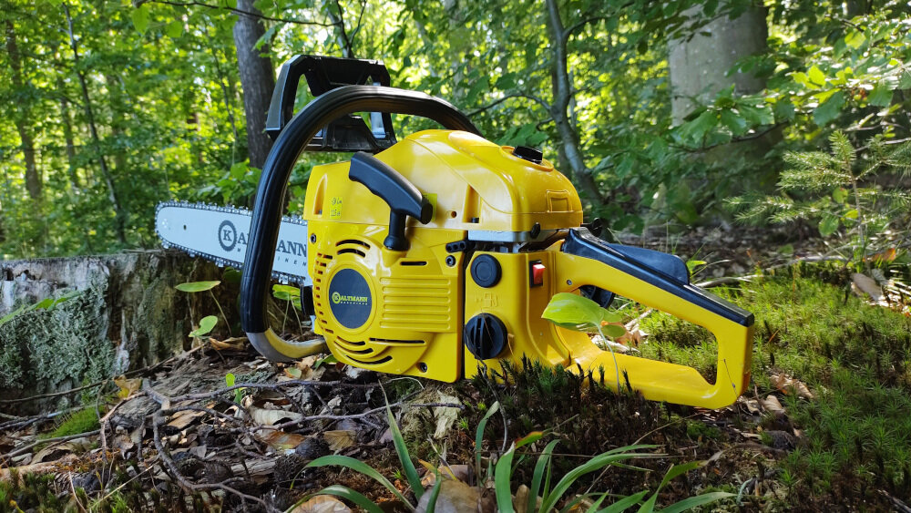 Żółta piła spalinowa leży na ziemi w lesie, z widocznym logo \'KALTMANN\' na prowadnicy. W tle znajdują się drzewa i roślinność leśna.
