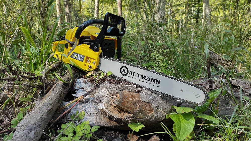Żółta piła spalinowa leży na ściętym pniu w lesie. Na prowadnicy widoczny jest napis \'KALTMANN Maschinen\'.