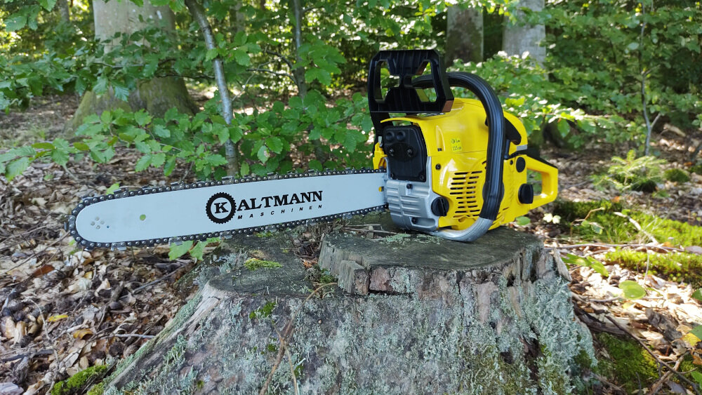 Żółta piła spalinowa leży na pniu drzewa w lesie. Na prowadnicy widoczny jest napis \'KALTMANN MASCHINEN\'.