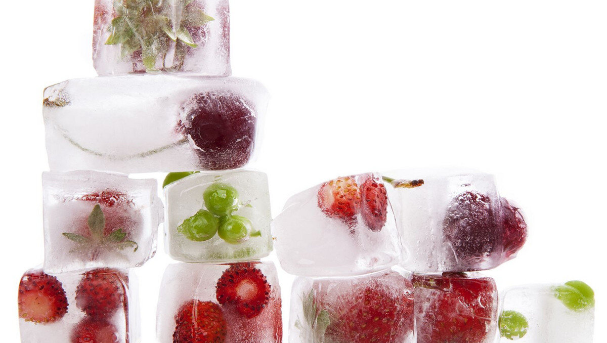 Kostki lodu z zamrożonymi owocami, takimi jak truskawki, winogrona i wiśnie, ułożone w stos. Tło jest białe, co podkreśla przezroczystość lodu.