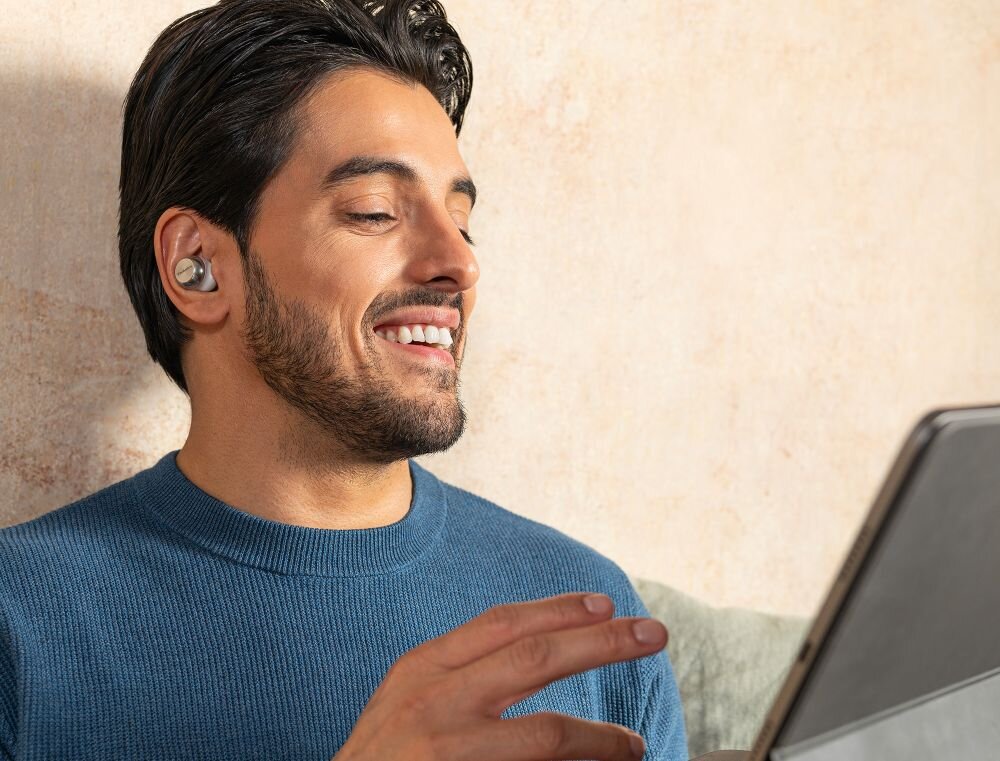 Osoba nosi srebrne słuchawki dokanałowe i patrzy na ekran laptopa. Tło jest jasne, a osoba ma na sobie niebieski sweter.