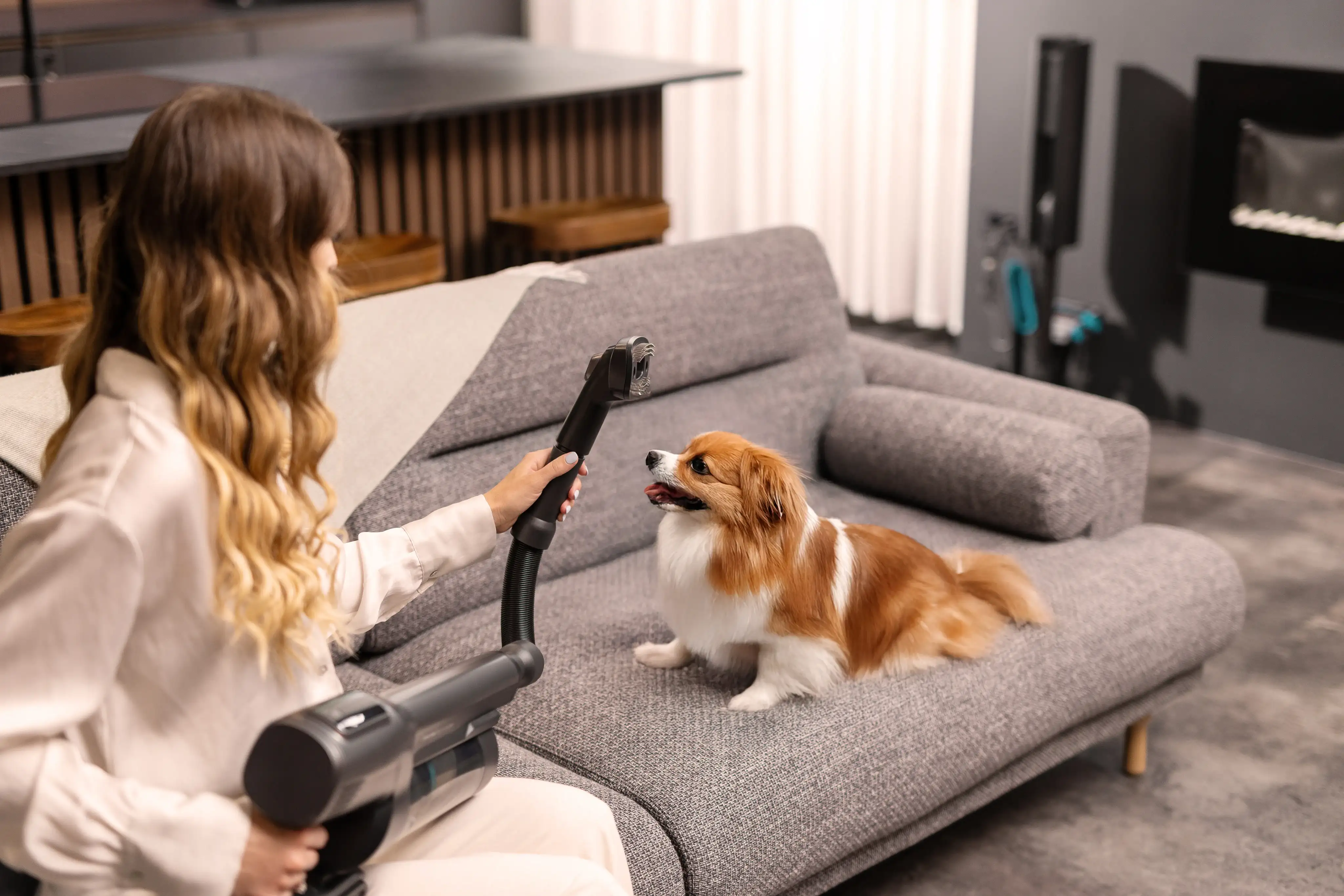 Osoba trzyma odkurzacz, siedząc na szarej kanapie, obok której znajduje się mały pies. W tle widoczny jest nowoczesny wystrój wnętrza z zasłonami i kominkiem.