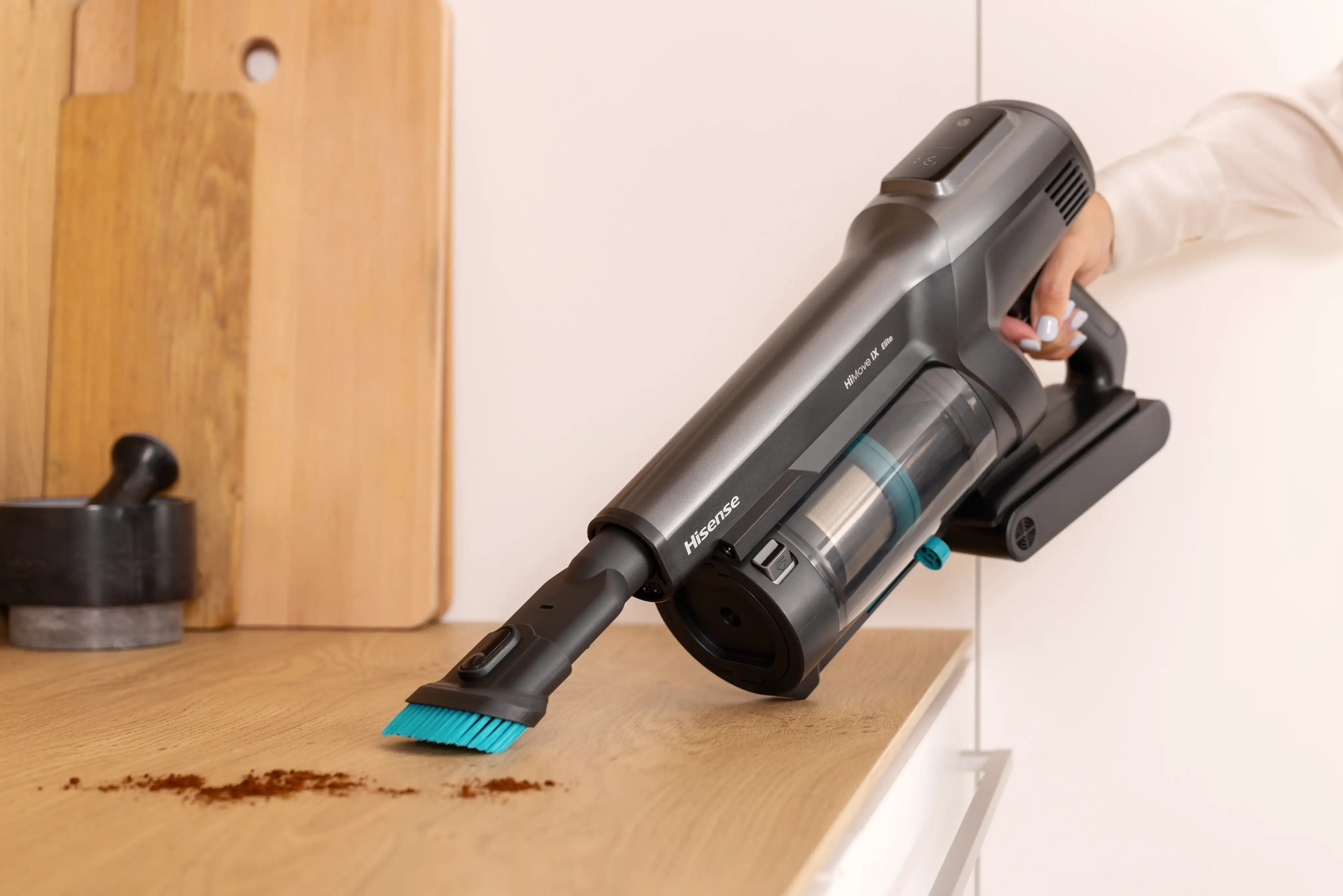 Osoba używa odkurzacza ręcznego Hisense HiMove IX do czyszczenia blatu z rozsypanej kawy. Na odkurzaczu widoczne są napisy \'Hisense\' i \'HiMove IX\'.