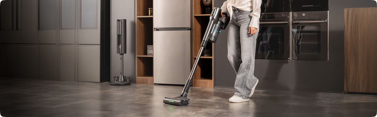 Osoba używa odkurzacza pionowego na podłodze w nowoczesnej kuchni. W tle widoczne są szafki kuchenne, lodówka i piekarnik.