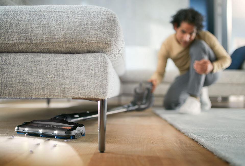 Odkurzacz mopujący znajduje się pod kanapą na drewnianej podłodze, zbierając drobne zanieczyszczenia. W tle widoczna jest osoba klęcząca na dywanie.