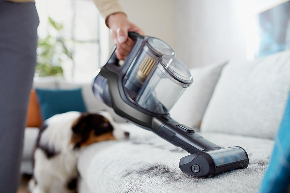 Osoba używa odkurzacza ręcznego na kanapie, obok leży pies. Odkurzacz ma przezroczysty pojemnik na kurz i ciemną obudowę.