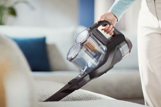Osoba trzyma odkurzacz ręczny z przezroczystym pojemnikiem na kurz, używając go do czyszczenia powierzchni mebla. Odkurzacz ma nowoczesny design i ciemną kolorystykę.