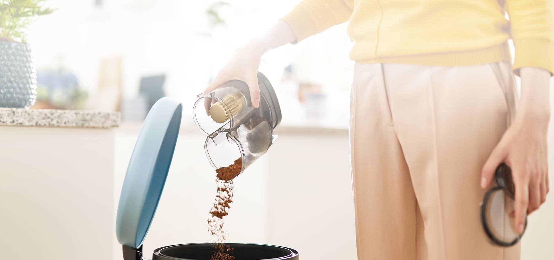 Osoba opróżnia pojemnik z odkurzacza do kosza na śmieci. W tle widoczne jest jasne wnętrze kuchni.